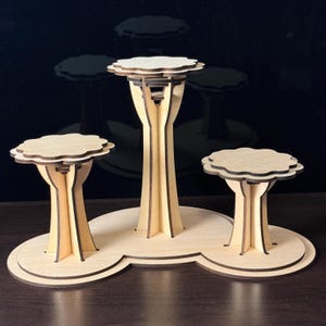May include: A three-tiered wooden display stand with a natural finish. The stand features three platforms with a scalloped edge, supported by tree-like structures. The base is a connected, rounded shape.