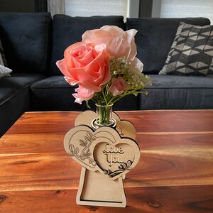 Op de afbeelding: Een houten vaas met twee hartvormige uitsnijdingen. De vaas heeft de woorden "Love You" gegraveerd op een van de harten. De vaas bevat een boeket roze rozen en witte bloemen.