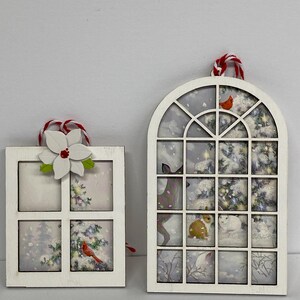 May include: Two white wooden Christmas ornaments shaped like windows with winter scenes inside. The larger ornament has a deer, a rabbit, and a bird in a snowy forest. The smaller ornament has a cardinal and a flower.