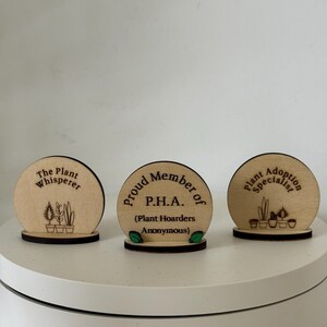 May include: Three wooden plant-themed desk signs. The signs are circular with a dark base. Text includes "The Plant Whisperer", "Proud Member of P.H.A. (Plant Hoarders Anonymous)", and "Plant Adoption Specialist". Each sign features plant illustrations.