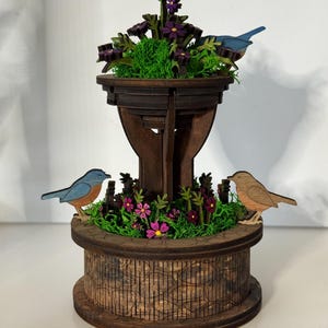 Puede incluir: Una escultura de fuente de madera de dos niveles con un acabado marrón oscuro. La estructura está decorada con musgo verde brillante, pequeñas flores moradas y rosas, y tres figuras de pájaros de madera en tonos azules y marrones.