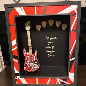 May include: A black shadow box with a red, white and black geometric pattern. Inside the box is a miniature electric guitar with a red, white and black paint scheme. The guitar is displayed with several guitar picks. The text "I'd pick you every single time." is written on the black background. The text "Guitar is not included" is written below the shadow box.
