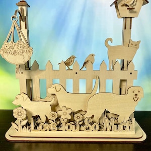 May include: A wooden decorative piece featuring a picket fence with cut-out dogs, cats, and birds. A hanging basket of flowers and a birdhouse are also included. The phrase "Love You So Much!" is cut out at the base. The piece is made of light-colored wood.