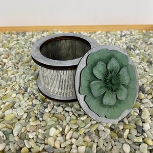 May include: A small wooden box with a white and black striped design. The lid is open and features a green flower with a raised design.