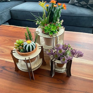 Puede incluir: Tres soportes de madera para plantas con patrones geométricos, cada uno con una planta suculenta en maceta diferente. Los soportes están dispuestos sobre una mesa de madera.