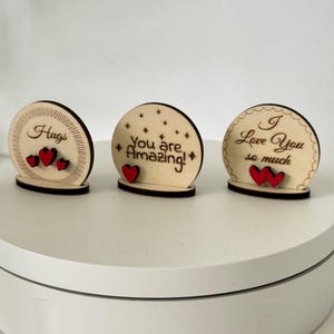 May include: Three wooden decorative signs with messages of love. The signs are round with a black base and feature the phrases "Hugs", "You are Amazing!", and "I Love You so much". Each sign has red heart accents.