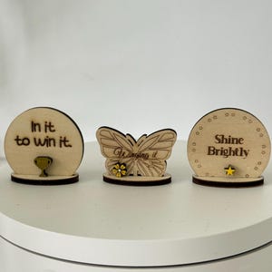 Op de afbeelding: Drie houten decoratieve stukken. Eén is een cirkel met de tekst "In it to win it" en een trofee. De tweede is een vlinder met de tekst "Winging it" en een bloem. De derde is een cirkel met de tekst "Shine Brightly" en een ster.