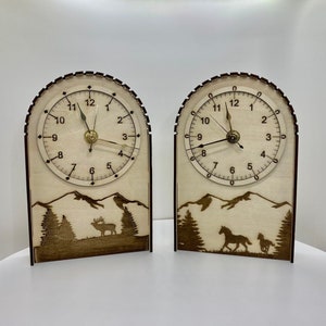 May include: Two wooden clocks with a mountain and animal silhouette design. The clocks have a white face with black numbers and hands. The clock on the left has a deer silhouette and the clock on the right has a horse silhouette.
