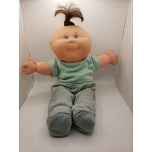 May include: A doll with a round, peach-toned face, blue eyes, and a single tuft of brown hair standing upright on its head. The doll wears a light green short-sleeved shirt and matching light green pants, with arms outstretched to the sides.