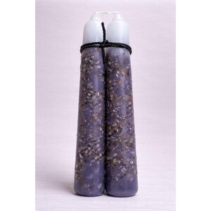 May include: Two purple taper candles bound together with a black rope. The candles have a textured surface with embedded flecks of gold and white. The tops of the candles are white.
