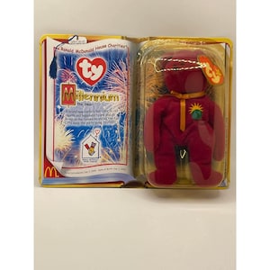 Puede incluir: Un oso de peluche rojo Millennium Bear con una etiqueta amarilla, dentro de una funda de plástico transparente. El embalaje incluye un folleto con el texto "Ronald McDonald House Charities" y "Millennium the Bear".