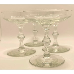May include: Four clear champagne coupe glasses with decorative stems. The glasses have a wide, shallow bowl and a decorative stem. The bases are round and the glasses are arranged on a white surface. The glasses are made of clear glass.