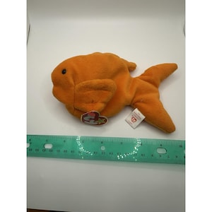 Può includere: Un pesce giocattolo di peluche arancione con un occhio nero e pinne. Il giocattolo ha un'etichetta attaccata con un logo a forma di cuore e il nome "Goldie". Un righello è posto accanto al giocattolo.