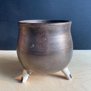 May include: A rounded ceramic planter with a metallic bronze finish and a smooth, curved body. The vessel is supported by three short, white, speckled legs that feature small black accents at the tips, providing a stable base for the pot.