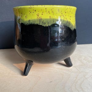 May include: A ceramic planter with three short legs, featuring a glossy black base and a bright yellow rim with dark brown speckles. The vessel has a rounded, bulbous body shape and a smooth, reflective finish on the exterior surface.