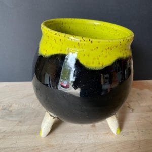 May include: A round ceramic pot with three short, off-white legs. The body of the pot is glossy black with a speckled texture, while the upper rim features a vibrant, bright yellow glaze that drips down slightly over the black surface.