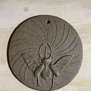May include: A round, brown ceramic wall hanging featuring a detailed relief of a beetle with outstretched wings. The beetle is centered, with radiating lines emanating from its head. A small hole is at the top for hanging.