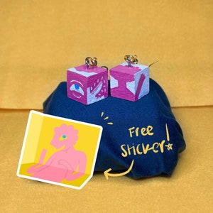 May include: Two pink and white painted wooden cube earrings with gold hooks. The earrings are on a blue fabric surface with the text "Free Sticker!" written in white. A sticker with a pink cartoon figure is also on the surface.