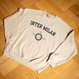 May include: A beige crewneck sweatshirt featuring the text "INTER MILAN" in black, arched across the top. Below, a circular emblem with "EST. 1908" and a star. The sweatshirt is made of a soft material.
