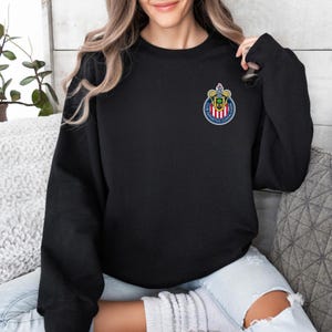 Op de afbeelding: Zwart sweatshirt met ronde hals en een klein, kleurrijk embleem op de linkerbovenborst. Het embleem is cirkelvormig met een wapenontwerp, inclusief rode, witte, blauwe en gouden elementen. Het sweatshirt is effen zwart.