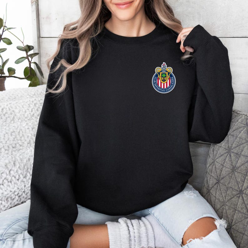 Op de afbeelding: Zwart sweatshirt met ronde hals en een klein, kleurrijk embleem op de linkerbovenborst. Het embleem is cirkelvormig met een wapenontwerp, inclusief rode, witte, blauwe en gouden elementen. Het sweatshirt is effen zwart.