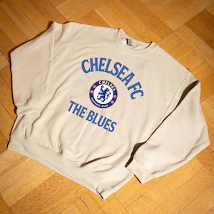 May include: A beige sweatshirt with the text "CHELSEA FC" in blue, arched above a circular emblem and the words "THE BLUES" below. The emblem features a blue lion and the words "CHELSEA FOOTBALL CLUB".