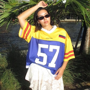 May include: A yellow and blue jersey with the number 57 in white. The jersey has red and yellow stripes on the sleeves and the text "PATRIA STUDIOS" across the chest. The jersey is worn over a white lace skirt.