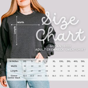 Op de afbeelding: Donkergrijs Gildan 18000 sweatshirt met ronde hals voor volwassenen met een maattabel. De tabel toont afmetingen in inches voor breedte, lengte en mouw middenachter voor maten XS tot 5XL. De woorden "Size Chart" staan in witte letters.
