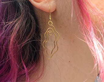 May include: Gold-colored wire earrings with a unique, abstract design. The earrings feature a flowing, organic shape with a spiral detail. The earrings are hanging from a person's ear, with pink and purple hair in the background.