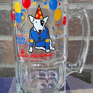 May include: Clear glass beer mug with a handle, decorated with colorful balloons and confetti. Features a cartoon Bull Terrier wearing a party hat and a blue sweater with the word "Spuds". The Bud Light logo is also visible.