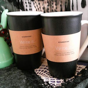 May include: Two matte black ceramic Starbucks mugs with white interiors and a white handle on the right mug. Each mug features a tan paper sleeve printed with the text Starbucks, Write it, and Create your own mug, Créez votre propre tasse.