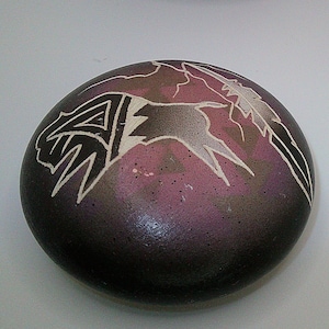 May include: A dark purple, oval stone with a carved design. The design features white and black geometric shapes and a bird-like figure. The stone has a smooth, polished surface and a gradient of purple and brown.