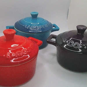 May include: Three small, ceramic lidded pots in red, teal, and dark purple. Each pot has a handle on either side and a knob on the lid. The word "AGOS" is visible on the teal pot, and "TACOS" and "GOS" are visible on the other two.