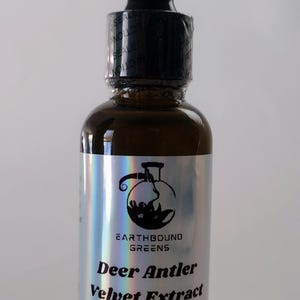 以下が含まれることがあります： 黒いドロッパーキャップが付いた茶色のガラス瓶。ラベルには「EARTHBOUND GREENS Deer Antler Velvet Extract 1 oz」と書かれています。ラベルは銀色のホログラフィック仕上げです。ボトルには透明な液体が入っています。