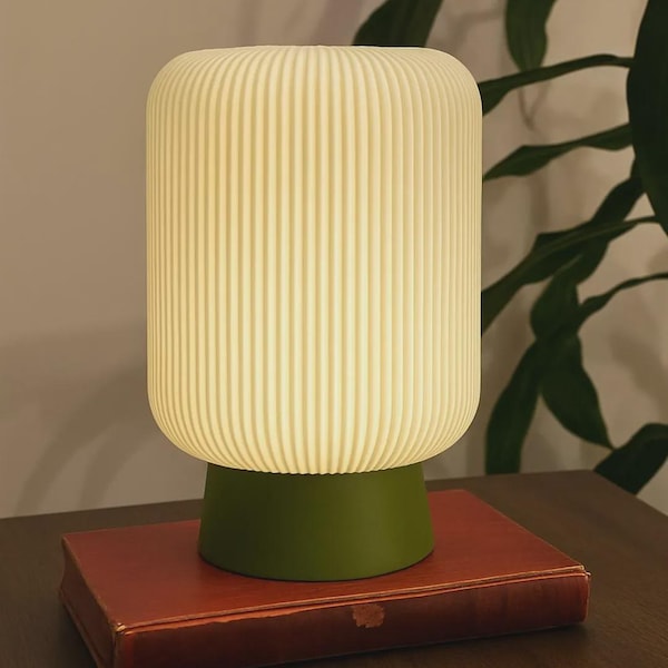 May include: A cylindrical table lamp with a ribbed, off-white translucent shade and a solid olive green base. The lamp is illuminated, emitting a soft glow, and is positioned on top of a rectangular, reddish-brown hardcover book.