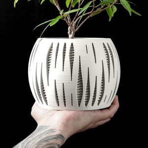 Peut inclure: Un pot de fleurs sphérique blanc avec un design découpé unique, contenant une petite plante verte. La surface du pot présente des ouvertures verticales en forme de goutte d'eau. La plante a des feuilles vertes et des tiges brunes. Le pot est tenu dans une main sur fond noir.