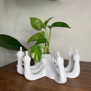 May include: White ceramic planter shaped like an octopus with all of its tentacles giving the middle finger. A green potted plant is in the octopus's head.