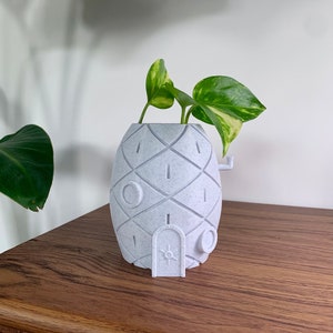 Puede incluir: Una maceta en forma de piña azul claro con una puerta y detalles circulares. La maceta contiene una planta de hojas verdes y amarillas. La maceta está sobre una superficie de madera.