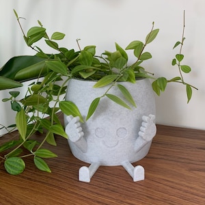 Peut inclure: Un pot en céramique gris en forme de personne souriante avec des bras et des jambes. Le pot est rempli d'une plante verte feuillue.
