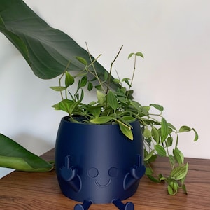 May include: A matte navy blue cylindrical planter featuring a molded character with a smiling face, two legs extending from the base, and two arms raised with fingers extended in a gesture, containing a green trailing plant with pointed leaves.