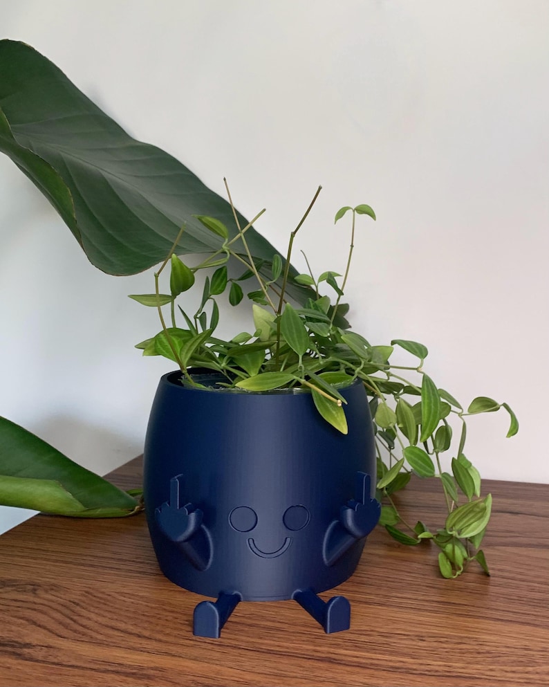 May include: A matte navy blue cylindrical planter featuring a molded character with a smiling face, two legs extending from the base, and two arms raised with fingers extended in a gesture, containing a green trailing plant with pointed leaves.