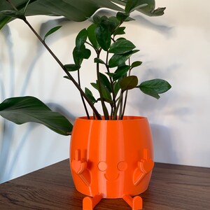 May include: A bright orange cylindrical planter featuring a molded character design with a smiling face, two legs extending from the base, and two arms raised with fingers extended in a gesture, containing a green Zamioculcas zamiifolia plant.