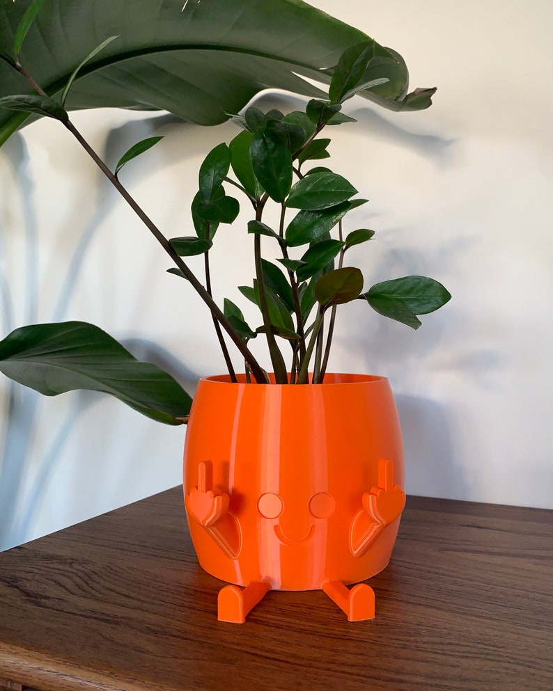 May include: A bright orange cylindrical planter featuring a molded character design with a smiling face, two legs extending from the base, and two arms raised with fingers extended in a gesture, containing a green Zamioculcas zamiifolia plant.