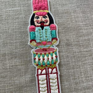 May include: Embroidered patch of a toy soldier nutcracker in a pink hat and teal jacket. The nutcracker has a black mustache and is holding drumsticks. A red drum with gold trim and white accents is at the bottom of the patch.