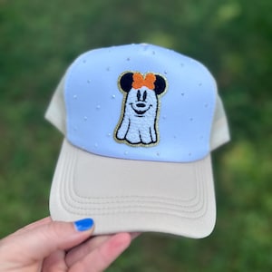 May include: A light blue and tan trucker hat with a cartoon ghost design. The ghost has a Minnie Mouse bow and is outlined in gold. The hat has small, sparkly embellishments on the front panel.