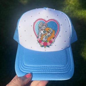 May include: A light blue and white trucker hat featuring a central embroidered patch of two cartoon dogs inside a pink heart frame with floral accents. The white front panel is decorated with small scattered rhinestones, and the brim is solid blue.