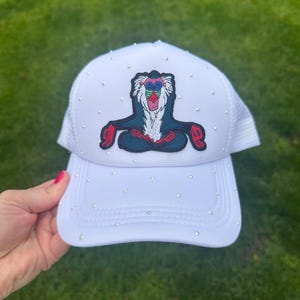 May include: White trucker hat with a mesh back and a curved brim. The front of the hat features a colorful embroidered image of a cartoon baboon. The hat is embellished with small, sparkling rhinestones.
