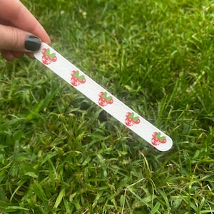 May include: A white nail file featuring a repeating pattern of red circular ornaments with white snowflake details and green bows. The nail file has a textured surface and is long and narrow in shape, designed for grooming fingernails.