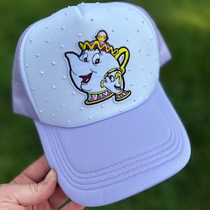 May include: A lavender trucker hat featuring an embroidered image of Mrs. Potts and Chip from Beauty and the Beast. The front panel is embellished with small, sparkling rhinestones. The hat has a curved brim and a mesh back.