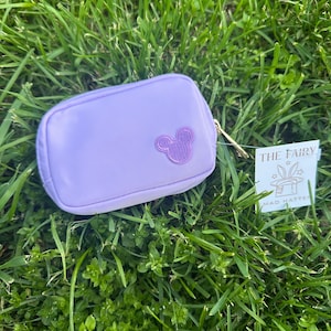 May include: A small, lavender-colored pouch with a gold zipper and a Mickey Mouse silhouette on the front. The pouch is set against a backdrop of green grass. A tag that reads "THE FAIRY MAD HATTER" is also visible.